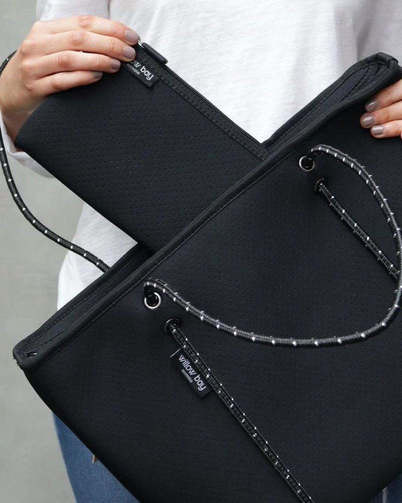 Woman holding a black perforated neoprene tote bag with contrast rope handles and a matching zip pouch.