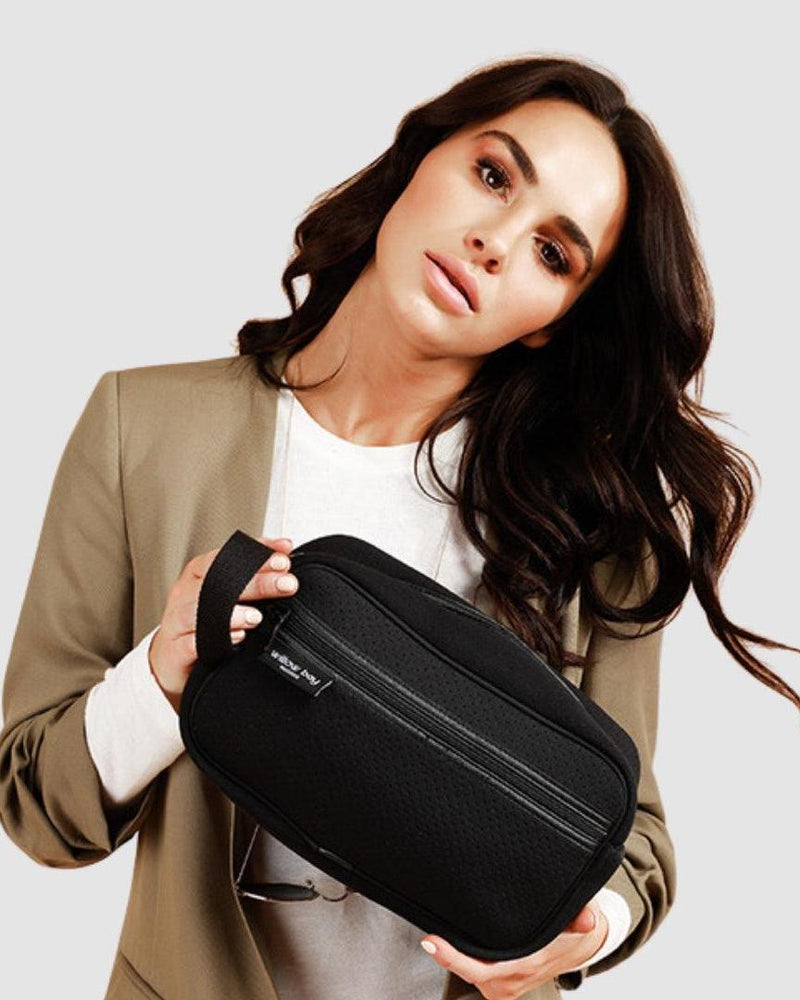 Woman holding a black neoprene cosmetic bag with a wrist strap and front zip pocket.