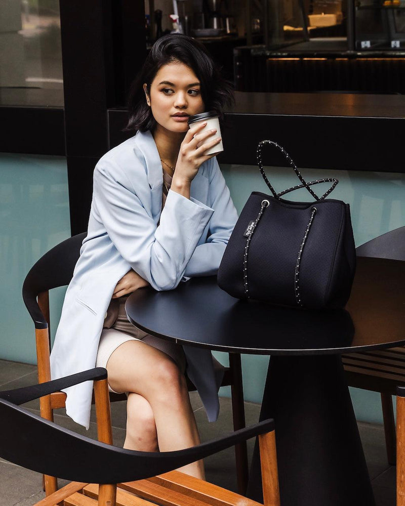 Woman with a black perforated neoprene Daydreamer Mini Tote, featuring studded details and a rope handle, at a cafe.
