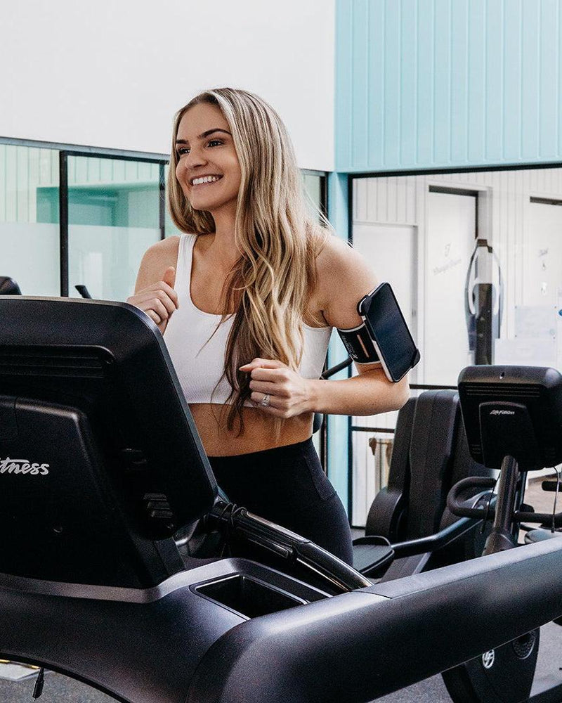 Smiling woman running on a treadmill, wearing a white sports bra and black Re-Active Sport Phone Holder on her left arm.