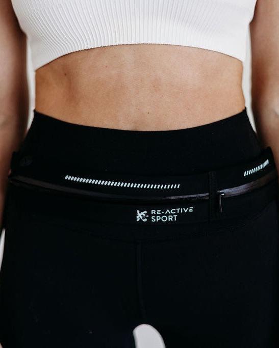 Black Re-Active Sport running belt with white logo, zipper, and reflective stripe worn on a person's waist.